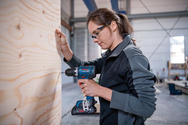 GSR 18V-90 C Một người mặc đồ bảo hộ đang khoan vào tấm gỗ trong xưởng.