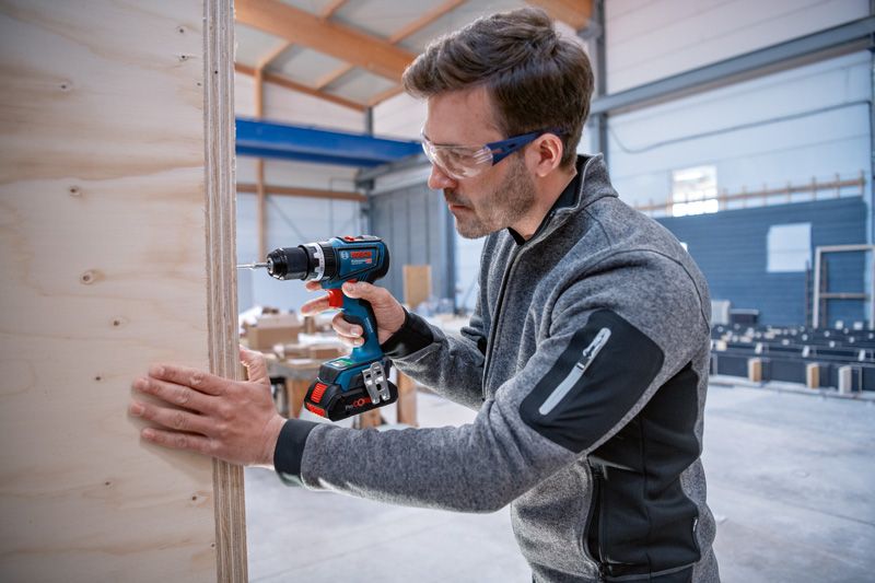 GSB 18V-90 C Người mặc đồ bảo hộ đang khoan vào tấm ván gỗ bên trong xưởng.