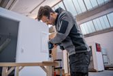 Một người mặc đồ bảo hộ đang khoan lỗ trên gỗ bằng máy khoan điện không dây.