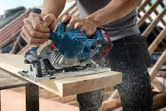 Một người đang cắt một tấm ván gỗ bằng máy cưa tròn không dây, làm tung tóe mùn cưa.
