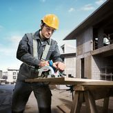 Một người mặc đồ bảo hộ đang cắt gỗ bằng máy cưa tròn không dây tại một công trường xây dựng.
