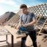 Một người mặc đồ bảo hộ đang cắt gỗ bằng máy cưa tròn không dây tại một công trường xây dựng.