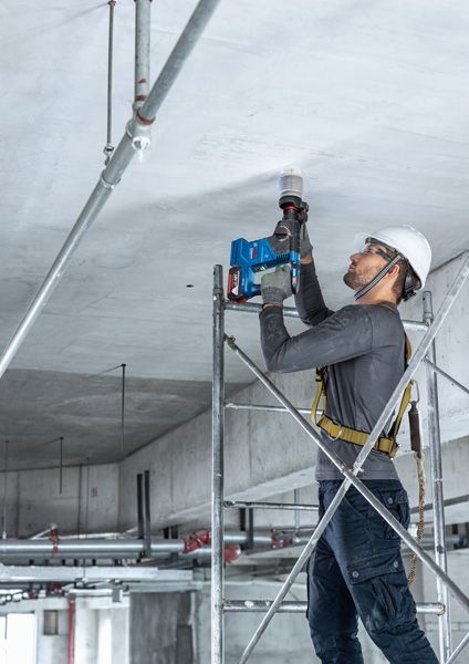 Một người mặc thiết bị an toàn đang khoan vào trần bê tông trong khi đứng trên giàn giáo.