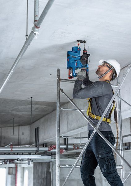 Một người mặc thiết bị an toàn đang khoan vào trần bê tông trong khi đứng trên giàn giáo.