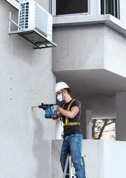 GBH 187-LI Một người mặc đồ bảo hộ đang khoan vào bức tường bê tông bên cạnh một thiết bị điều hòa không khí.