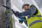 Một người mặc đồ bảo hộ đang cắt một ống kim loại bằng cưa lọng.