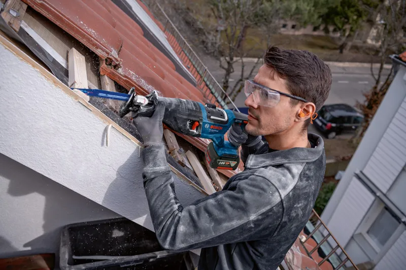 GSA 185-LI Một người mặc đồ bảo hộ đang sử dụng máy cưa lọng để cắt gỗ trên mái nhà.