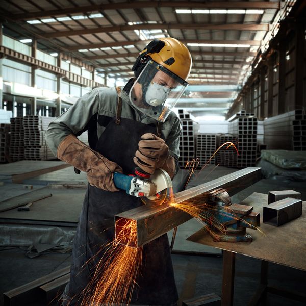 Một người mặc đồ bảo hộ đang mài một thanh kim loại bằng máy mài góc, tia lửa bắn ra.