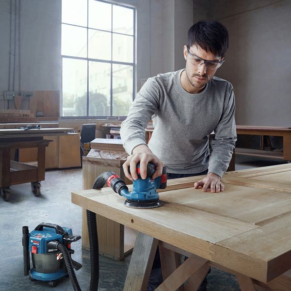 Một người mặc đồ bảo hộ đang chà nhám cửa gỗ bằng máy chà nhám quỹ đạo được kết nối.