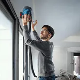 Người mặc đồ bảo hộ đang chà nhám trần nhà gần cửa sổ bằng máy chà nhám điện.