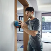 Một người mặc đồ bảo hộ đang chà nhám tường bằng máy chà nhám không dây.