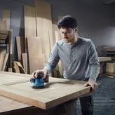 Người mặc đồ bảo hộ đang chà nhám cửa gỗ bằng máy chà nhám điện.