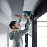 Một người mặc đồ bảo hộ đang chà nhám mép trần nhà bằng máy chà nhám điện.