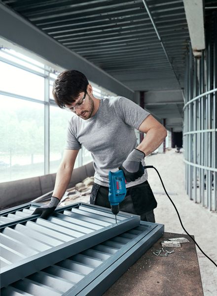 Người mặc đồ bảo hộ đang khoan vít vào khung kim loại tại công trường xây dựng.