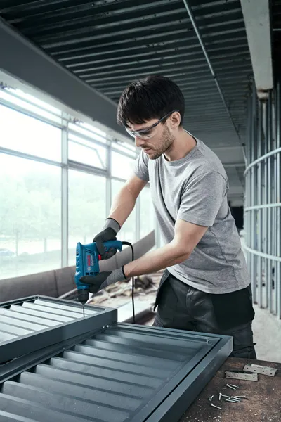 Một người mặc đồ bảo hộ sử dụng máy khoan điện để lắp ráp khung kim loại.