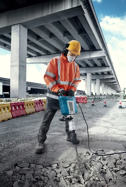 Một người mặc đồ bảo hộ đang phá bê tông bằng máy cắt điện lớn ở ngoài trời.