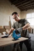 Một người mặc đồ bảo hộ đang sử dụng máy cưa lọng để cắt gỗ trên bàn làm việc.