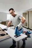 Một người mặc đồ bảo hộ đang cắt một tấm ván gỗ bằng máy cưa lọng trong xưởng.