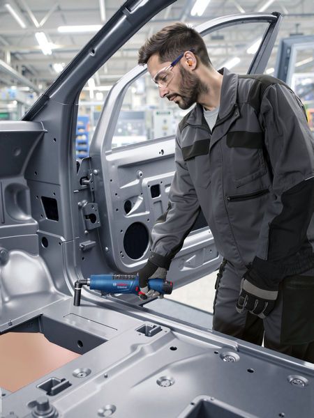 Người mặc thiết bị an toàn đang sử dụng dụng cụ điện trên khung ô tô trong môi trường công nghiệp.