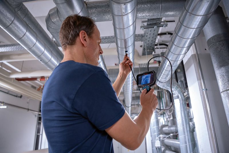 GIC 5-27 C Một người đang kiểm tra đường ống trên cao bằng camera kiểm tra trong môi trường công nghiệp.