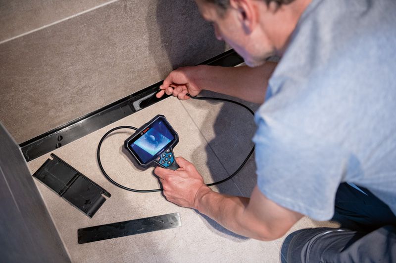 GIC 5-27 C Một người sử dụng camera kiểm tra để kiểm tra phía sau tấm bảng trên bức tường lát gạch.