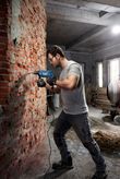 Một người mặc đồ bảo hộ đang khoan vào tường gạch bằng máy khoan tác động.
