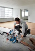 Một người mặc đồ bảo hộ đang cắt sàn gỗ bằng cưa góc.