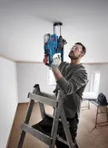 Một người mặc đồ bảo hộ đang khoan trần nhà trong khi đứng trên thang.