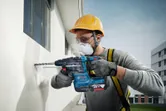 Một người mặc đồ bảo hộ đang khoan vào bức tường bên ngoài bằng một dụng cụ điện.