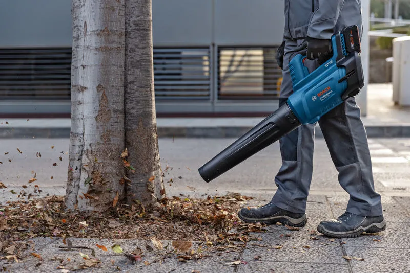 Một người mặc đồ bảo hộ sử dụng máy thổi lá không dây để thổi lá gần cây.