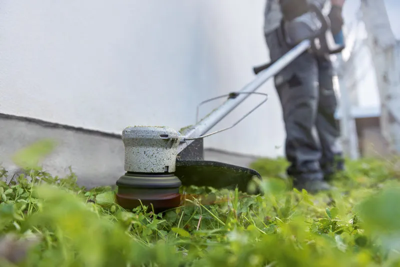 GRT 18V-33 Một người mặc đồ bảo hộ đang cắt cỏ dọc theo bức tường bằng máy cắt cỏ không dây.