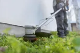 Một người mặc đồ bảo hộ đang cắt cỏ dọc theo bức tường bằng máy cắt cỏ không dây.