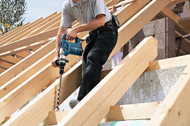 GBH 4-32 DFR Một người đang khoan vào dầm gỗ trên mái nhà mà không đeo thiết bị an toàn.