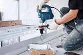 Một người mặc đồ bảo hộ đang khoan vào thanh kim loại bằng búa quay.