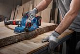 Một người mặc đồ bảo hộ đang bào một tấm ván gỗ bằng một dụng cụ không dây.