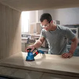 Một người mặc đồ bảo hộ đang chà nhám gỗ bằng máy chà nhám quỹ đạo ngẫu nhiên bên trong xưởng.