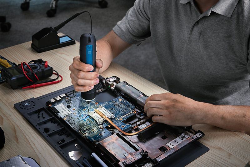 Người đang sử dụng tua vít không dây để lắp ráp máy tính xách tay trên bàn làm việc.