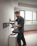 Người đeo thiết bị an toàn khoan vào tường để lắp giá đỡ bằng kim loại.