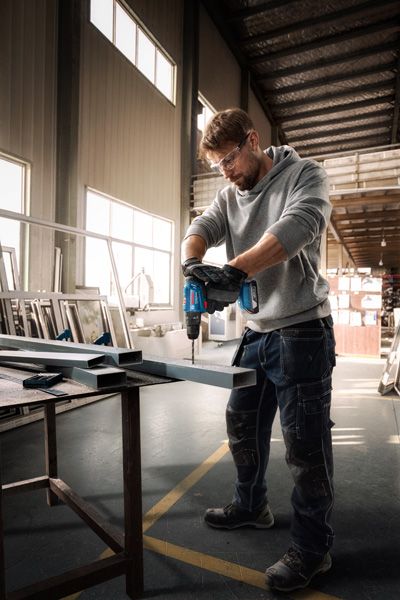 Một người mặc đồ bảo hộ đang khoan vào thanh dầm kim loại trong xưởng.