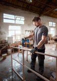 Một người mặc đồ bảo hộ đang khoan vào khung gỗ trong xưởng.