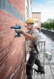 Một người mặc đồ bảo hộ đang khoan vào tường gạch trên giàn giáo.