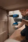 Một người đeo thiết bị an toàn đang lắp bản lề vào tủ gỗ bằng máy khoan không dây.