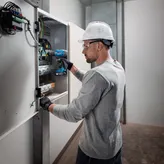 Một người mặc đồ bảo hộ đang sử dụng máy khoan không dây trên bảng điện.