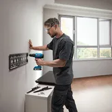 Một người mặc thiết bị an toàn đang khoan lỗ trên tường để gắn giá đỡ kim loại.