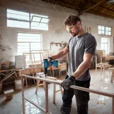 Một người mặc đồ bảo hộ đang khoan vào khung gỗ trong xưởng.