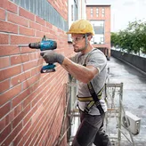 Một người mặc đồ bảo hộ đang khoan vào tường gạch trên giàn giáo.