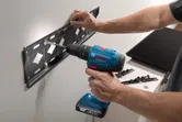 Một người đang khoan vít vào giá treo tường bằng máy khoan điện không dây.