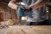 Người đeo thiết bị an toàn đang sử dụng máy khoan không dây để khoan lỗ trên tấm gỗ.