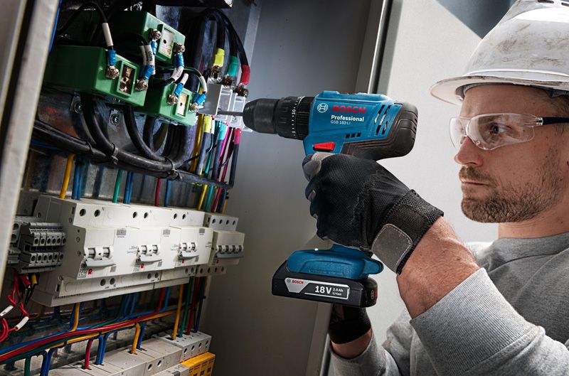Một người mặc đồ bảo hộ đang khoan vào bảng điều khiển đầy dây điện.