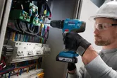 Một người mặc đồ bảo hộ đang khoan vào bảng điều khiển đầy dây điện.